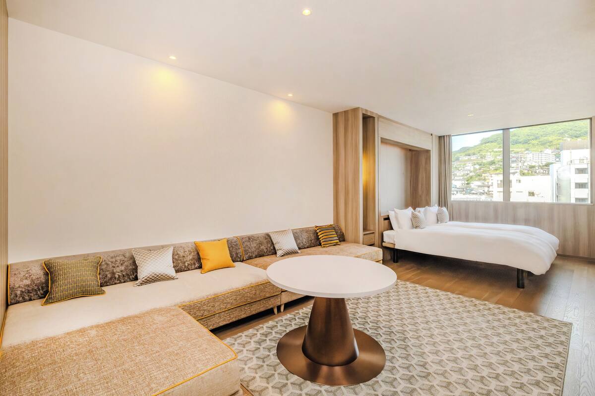 Ett modernt hotellrum med en soffa, ett runt bord, en säng och ett stort fönster med utsikt över staden.