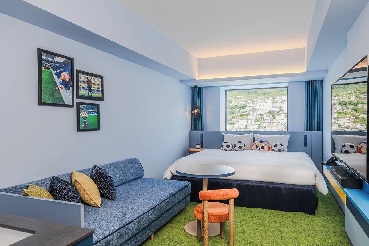 Ett hotellrum med en säng, en soffa, ett litet bord och en tv. Det finns tavlor på väggen och ett fönster med utsikt över byggnader.