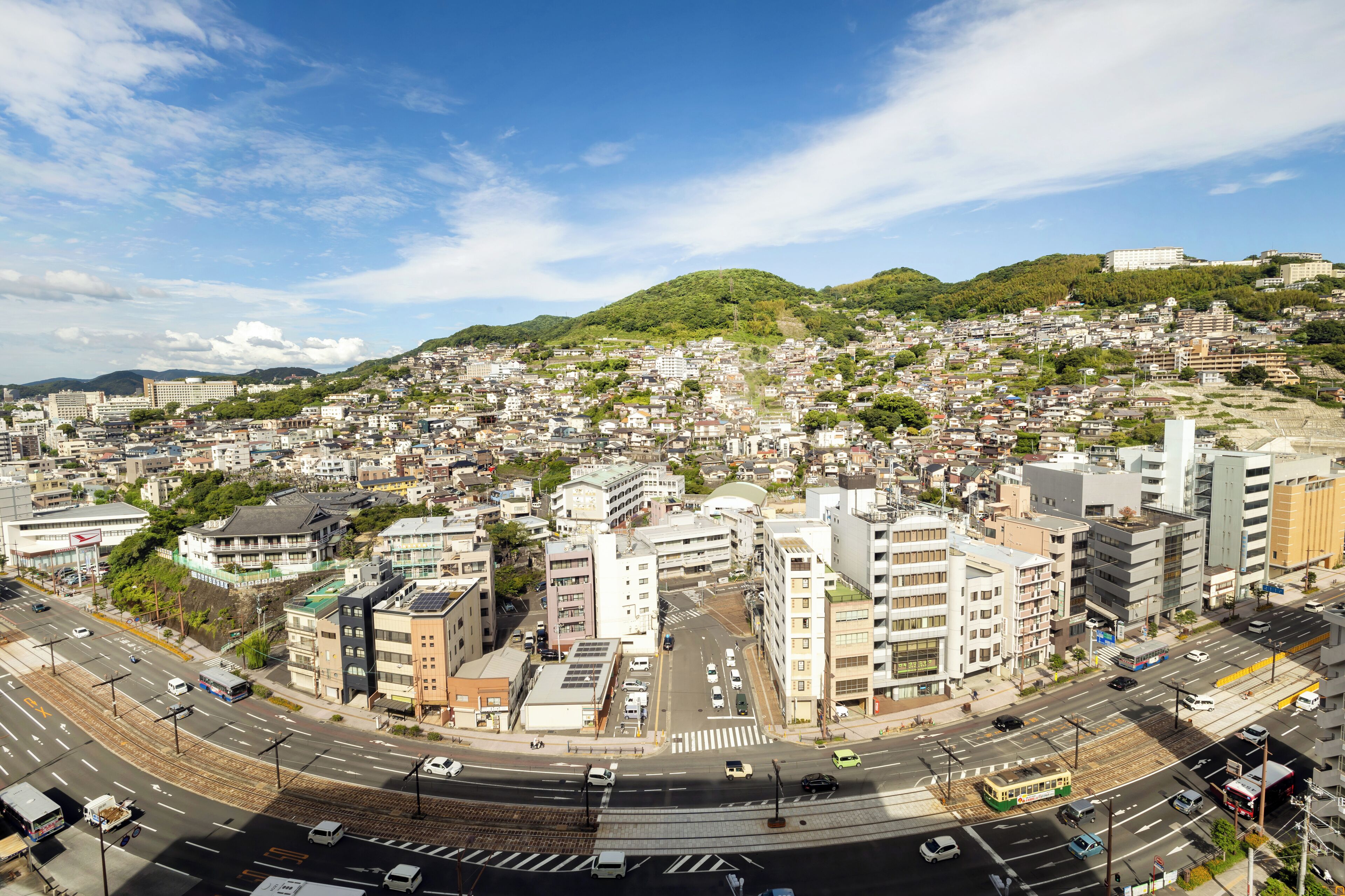 A panoramic view of a cityscape with a mix of residential and commercial buildings, a prominent hill with greenery, and a clear blue sky.