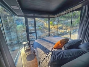 A modern room with a large bed, a small table, and a view of a rocky landscape through large windows. - Modern Glass Pod on Vineyard (Penticton)
