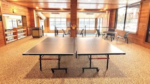 A room with two ping pong tables, wooden walls, a bookshelf, and a large window. - Ski-In/Ski-Out Condo | Hot Tub + Mountain Views (Lutsen)