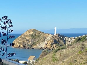 A coastal landscape with a lighthouse on a hill, a small house, and a clear blue sky. -   Stylish modern apartment with stunning lighthouse views, close to the beach (Castlepoint)