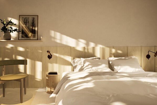 A bedroom with a bed, a chair, a bedside table, a wall-mounted lamp, and a framed picture.