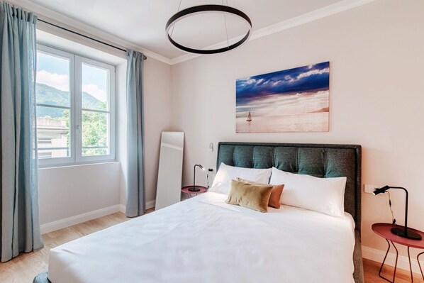 A bedroom with a bed, a painting of a beach scene, a window with curtains, and a bedside table with a lamp.