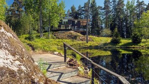 Una cabaña moderna con una terraza que da a un lago, rodeada de un bosque denso.