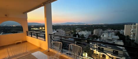 A balcony with a glass table and chairs, offering a view of a cityscape at dusk.