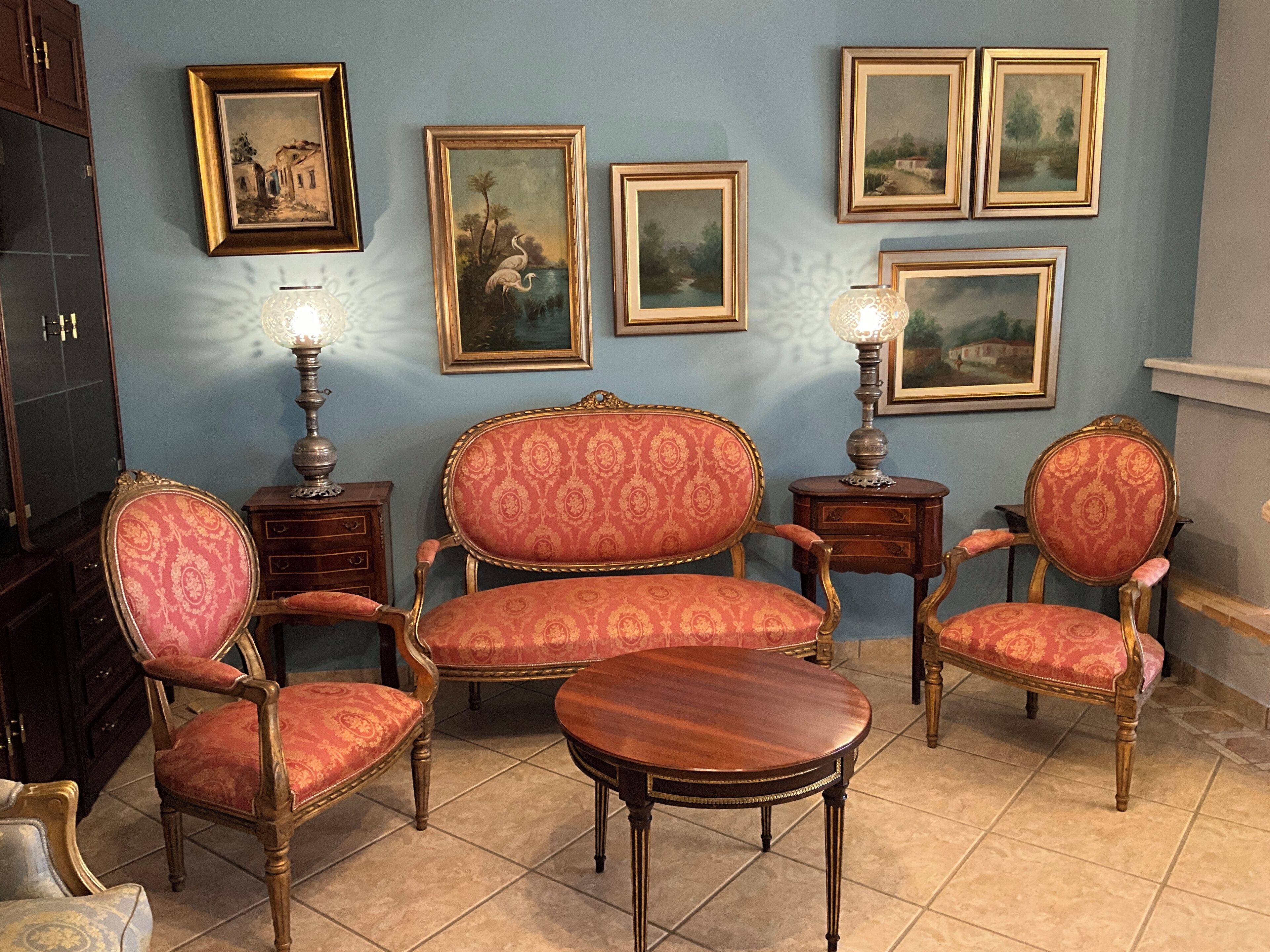 Une pièce avec un mur bleu, quatre tableaux encadrés et deux fauteuils en bois richement décorés, recouverts d’un tissu rouge.