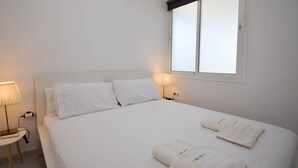 Cama bem arrumada com lençóis brancos e duas toalhas, uma luminária de cabeceira e uma janela com persianas.