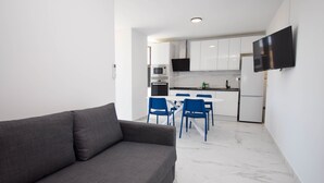 Uma sala moderna com sofá cinza, área de refeições com cadeiras azuis, cozinha com armários brancos e uma TV de tela plana na parede.