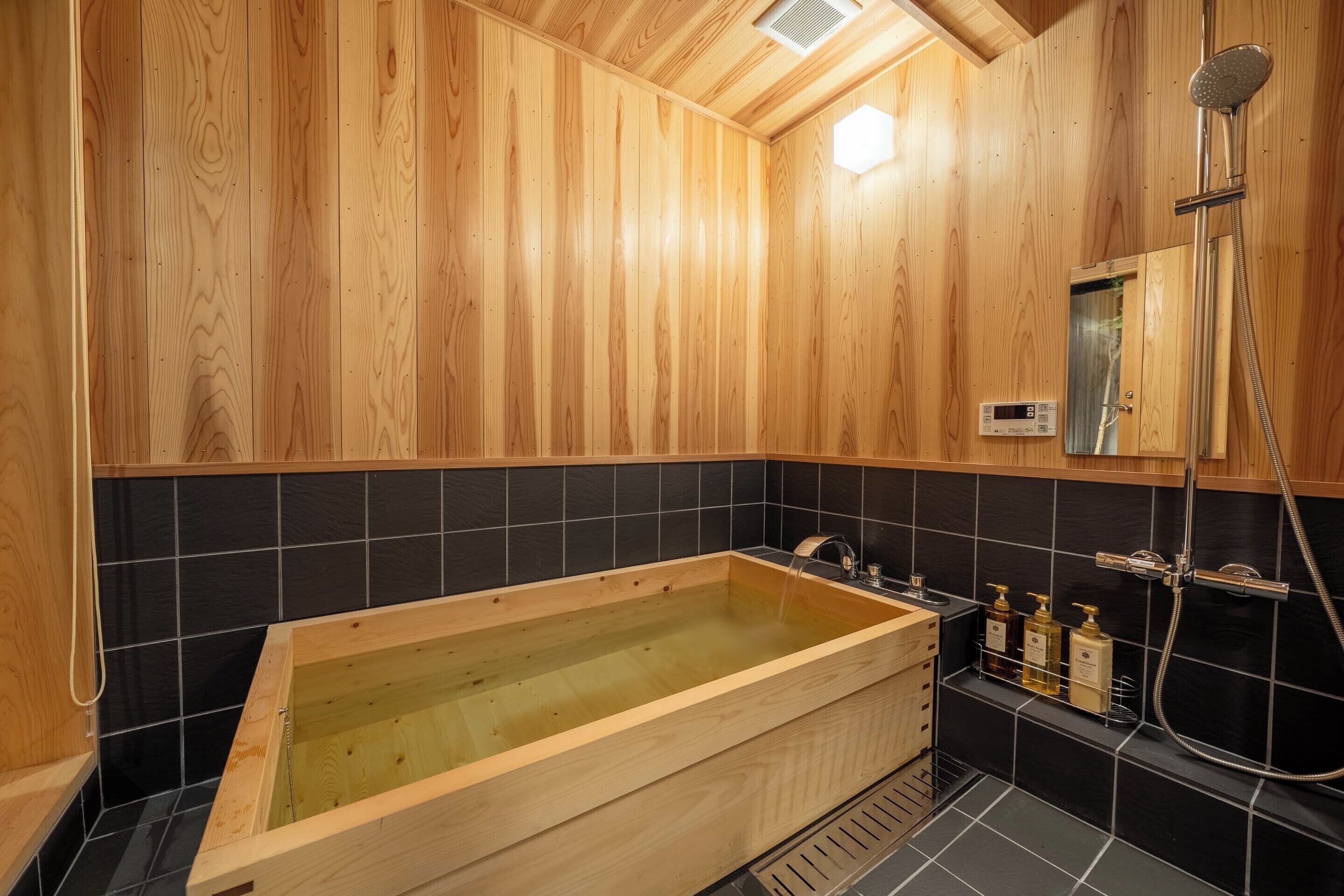 A wooden-paneled bathroom with a wooden bathtub, black tiled walls, and a shower area.