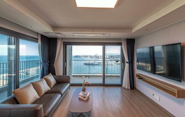 A modern hotel room with a sofa, a coffee table, a TV, and large windows offering a city and port view.