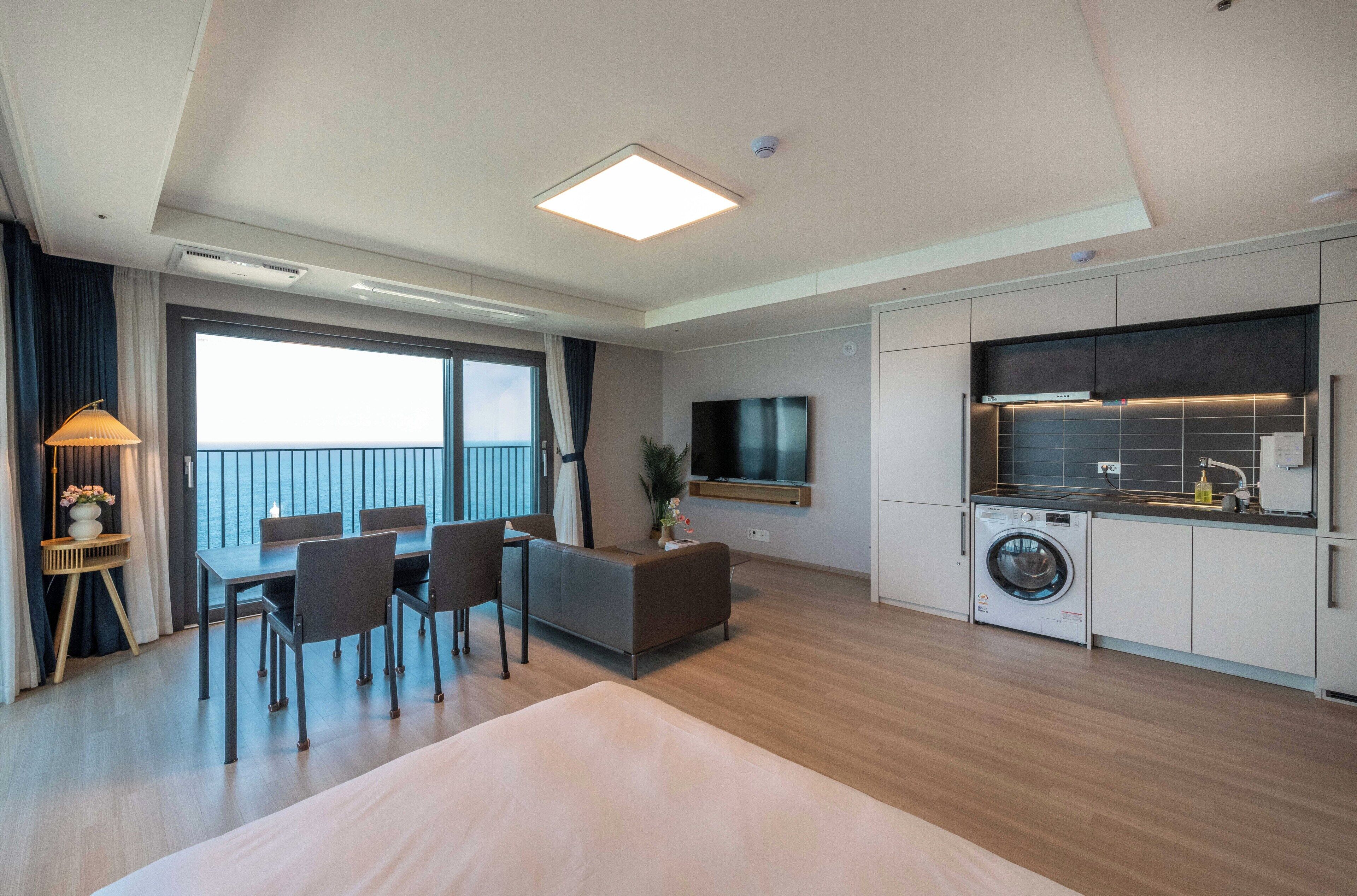 A modern hotel room with a large bed, a dining area with chairs, a TV, a washing machine, and a balcony with a view.