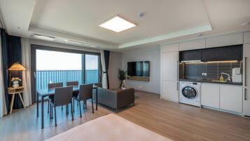 A modern hotel room with a large bed, a dining area with chairs, a TV, a washing machine, and a balcony with a view.