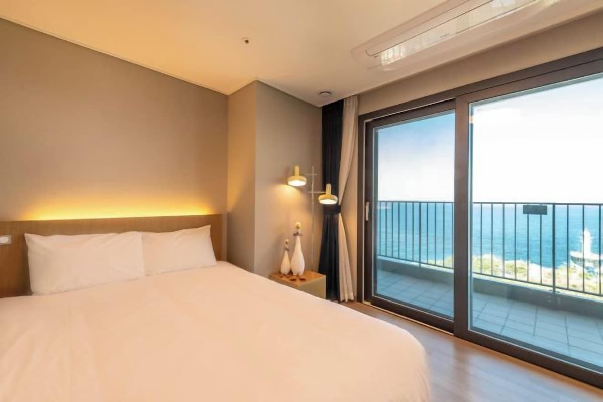 A hotel room with a large bed, a nightstand, a lamp, and a view of the ocean through a sliding glass door.