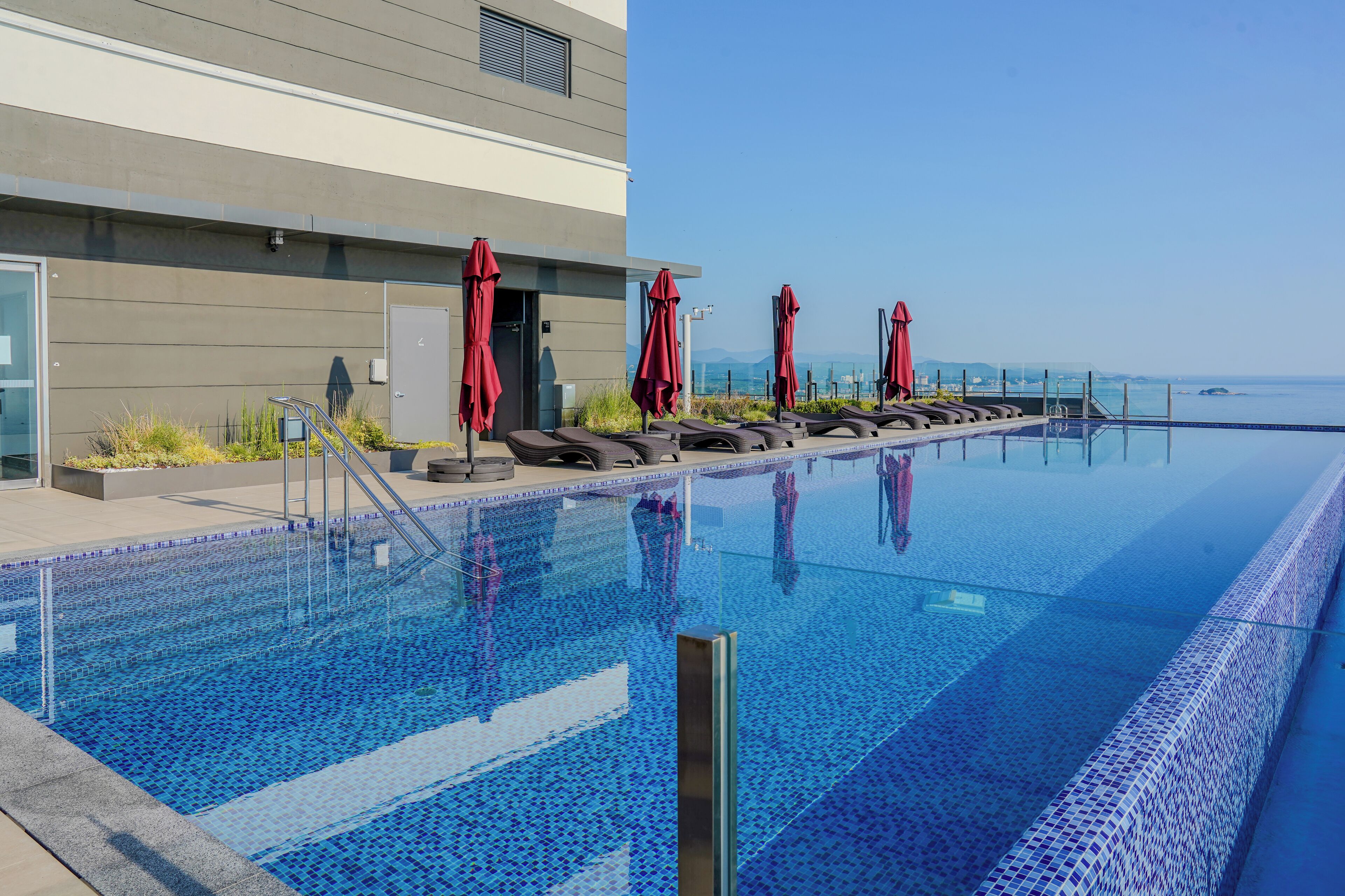 Un área de piscina moderna con sillones de descanso y sombrillas rojas, una vista despejada del mar y un edificio de fachada gris.