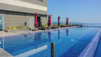 Un área de piscina moderna con sillones de descanso y sombrillas rojas, una vista despejada del mar y un edificio de fachada gris.