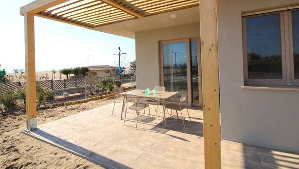 Un porxo cobert amb una taula, cadires, una porta de vidre i vista a la platja.