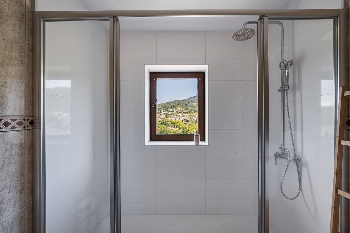 Une douche avec une porte en verre, une petite fenêtre donnant sur un paysage et un pommeau de douche.