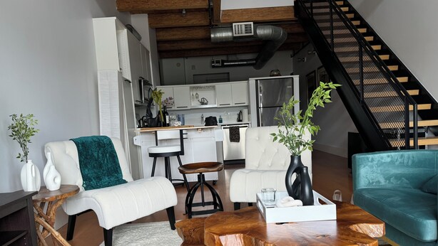 A modern living room with a sofa, a coffee table, and a teal couch. There is a kitchen in the background with white cabinets and a black staircase.