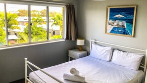 A neatly made bed with white linens and a rolled towel, a bedside lamp, and a painting of a boat on the wall. - U09 Tripcony 2 Bedroom w Balcony (Caloundra)