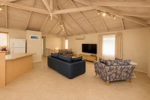 A spacious living area with a high ceiling, a flat-screen TV, a sofa, a patterned armchair, and a kitchen counter.