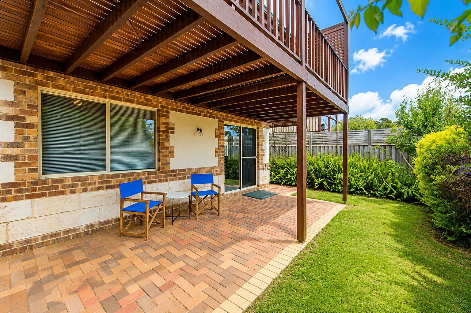 Une terrasse couverte avec un sol en briques, deux chaises bleues et une pergola en bois.