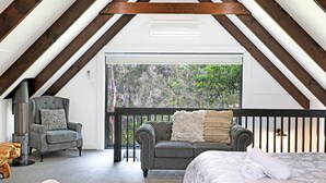 An attic room with a high ceiling, exposed wooden beams, a large sliding glass door leading to a balcony, a grey sofa, a chair, and a bed with a white duvet. - Waterfront Getaway, Pet Friendly - Jervis Bay - Book 3 Nights, Pay for 2 until 24 September 2026 (Saint Georges Basin)