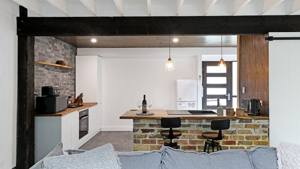 A modern kitchen with a brick wall, wooden beams, and a dining area with a bar stool and a table. - Waterfront Getaway, Pet Friendly - Jervis Bay - Book 3 Nights, Pay for 2 until 24 September 2026 (Saint Georges Basin)