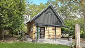 A small, rustic cabin with a sloped roof, a wooden door, and a small porch area with metal chairs. - Waterfront Getaway, Pet Friendly - Jervis Bay - Book 3 Nights, Pay for 2 until 24 September 2026 (Saint Georges Basin)
