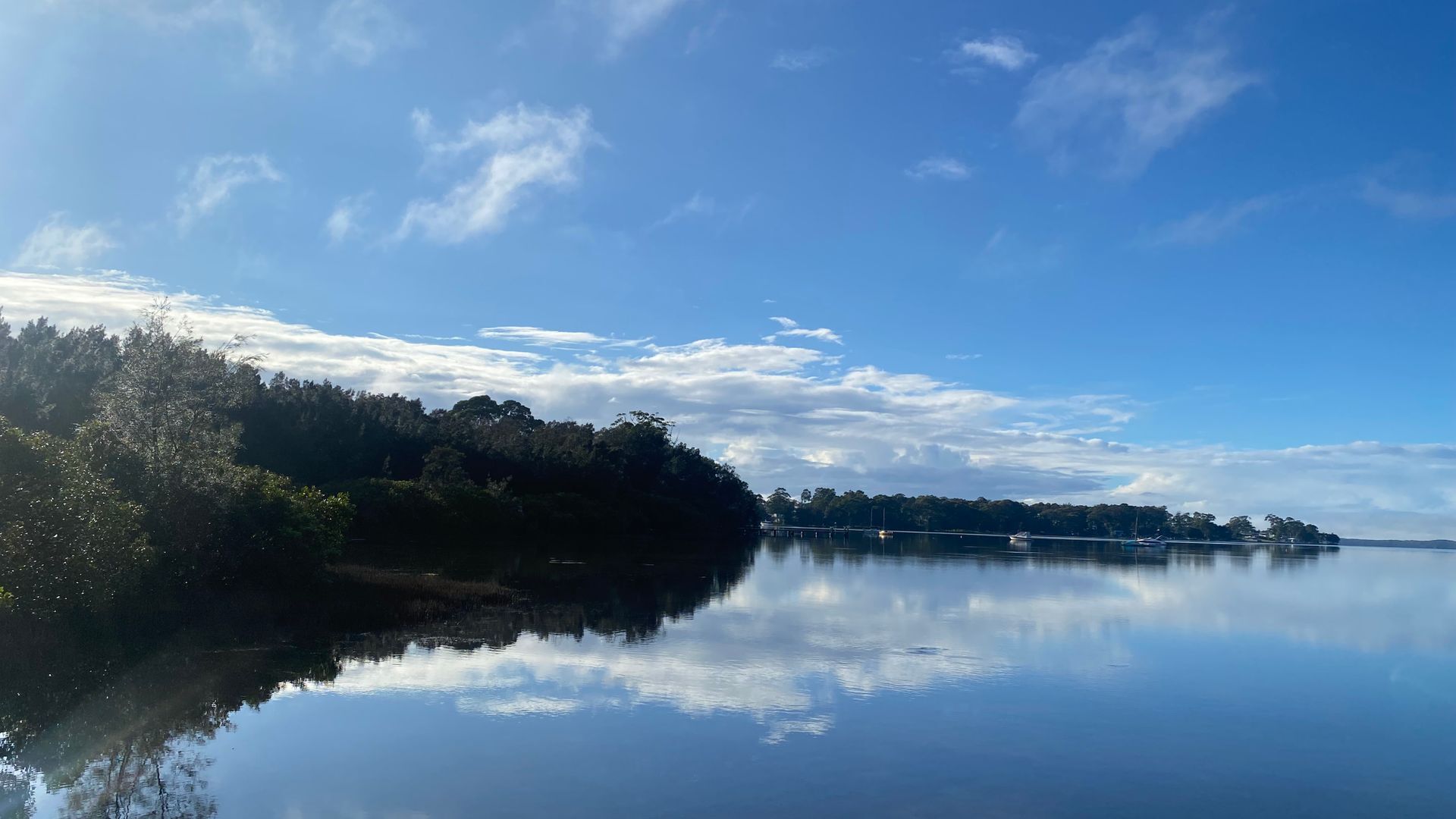 Um lago calmo com margens arborizadas e uma ponte ao longe.