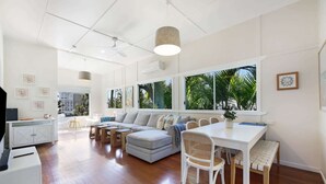 A spacious living area with a sofa, dining table, and chairs, large windows with a view of palm trees, and a ceiling fan. - 2-Bed Beach House in Burleigh Heads Amidst Nature (Burleigh Heads)