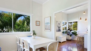 A dining area with a white table and chairs, a potted plant, and a view of palm trees through large windows. - 2-Bed Beach House in Burleigh Heads Amidst Nature (Burleigh Heads)