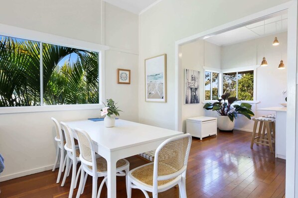 A dining area with a white table and chairs, a potted plant, and a view of palm trees through large windows.
