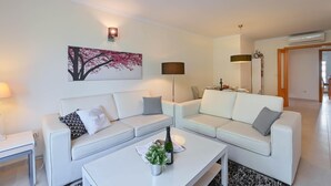 Un salon moderne avec deux canapés blancs, une table basse sur laquelle se trouvent une bouteille et des fleurs, une lampe et un tableau représentant un arbre, accroché au mur.