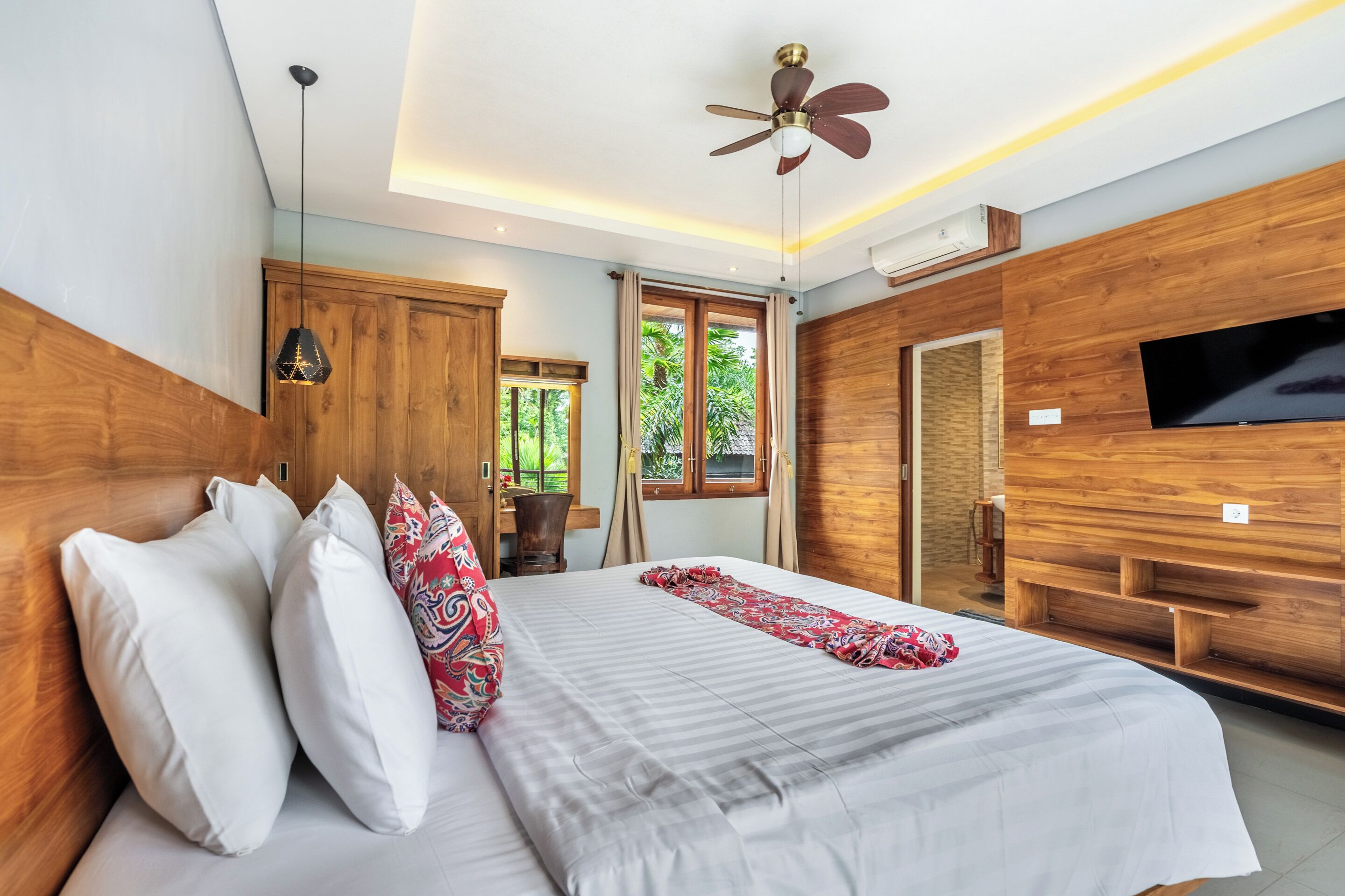 A modern bedroom with a large bed, a ceiling fan, a TV, and a window with a view of greenery.