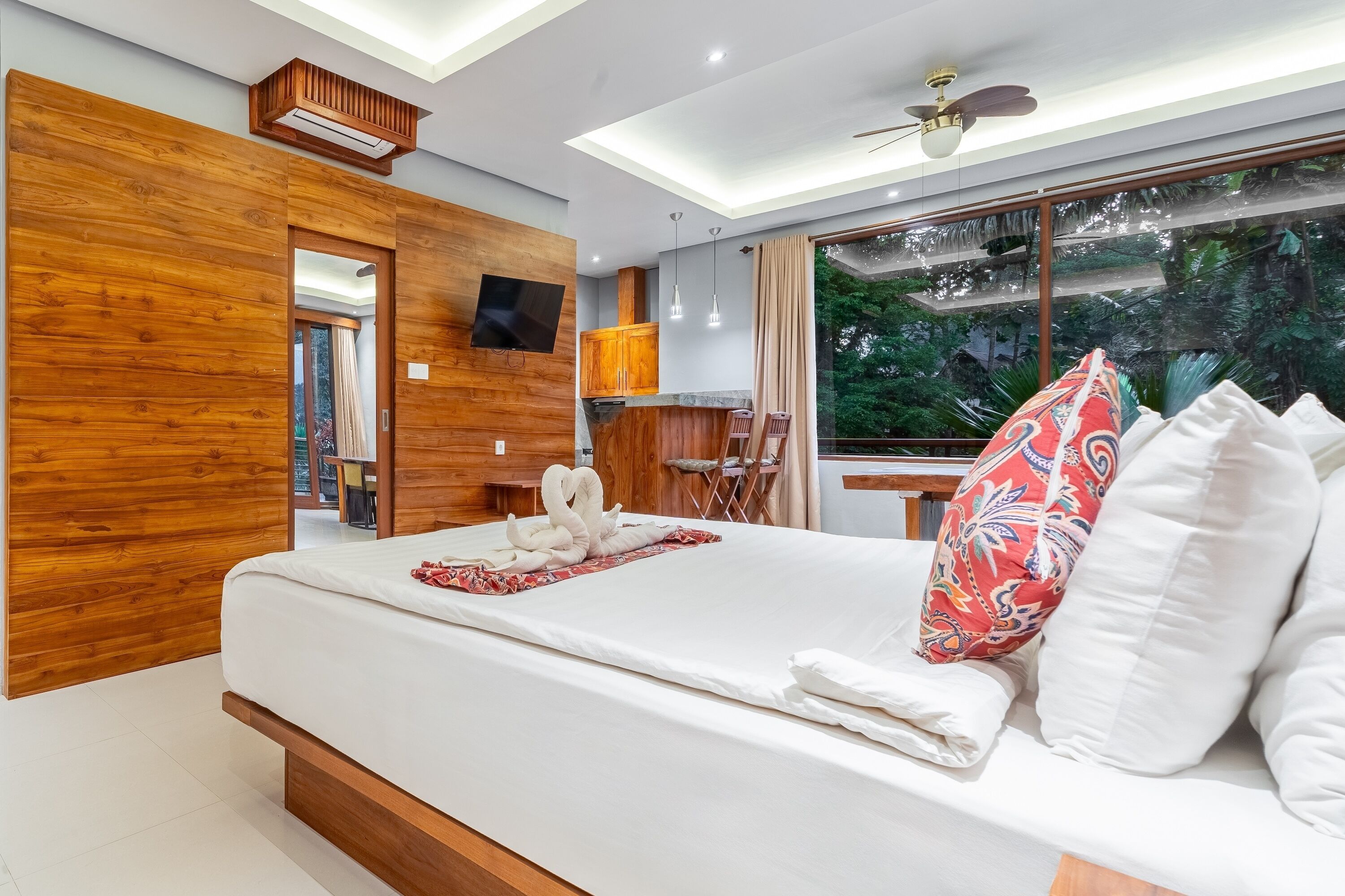 A modern hotel room with a wooden accent wall, a large bed with white linens, a TV mounted on the wall, and a view of lush greenery through the window.
