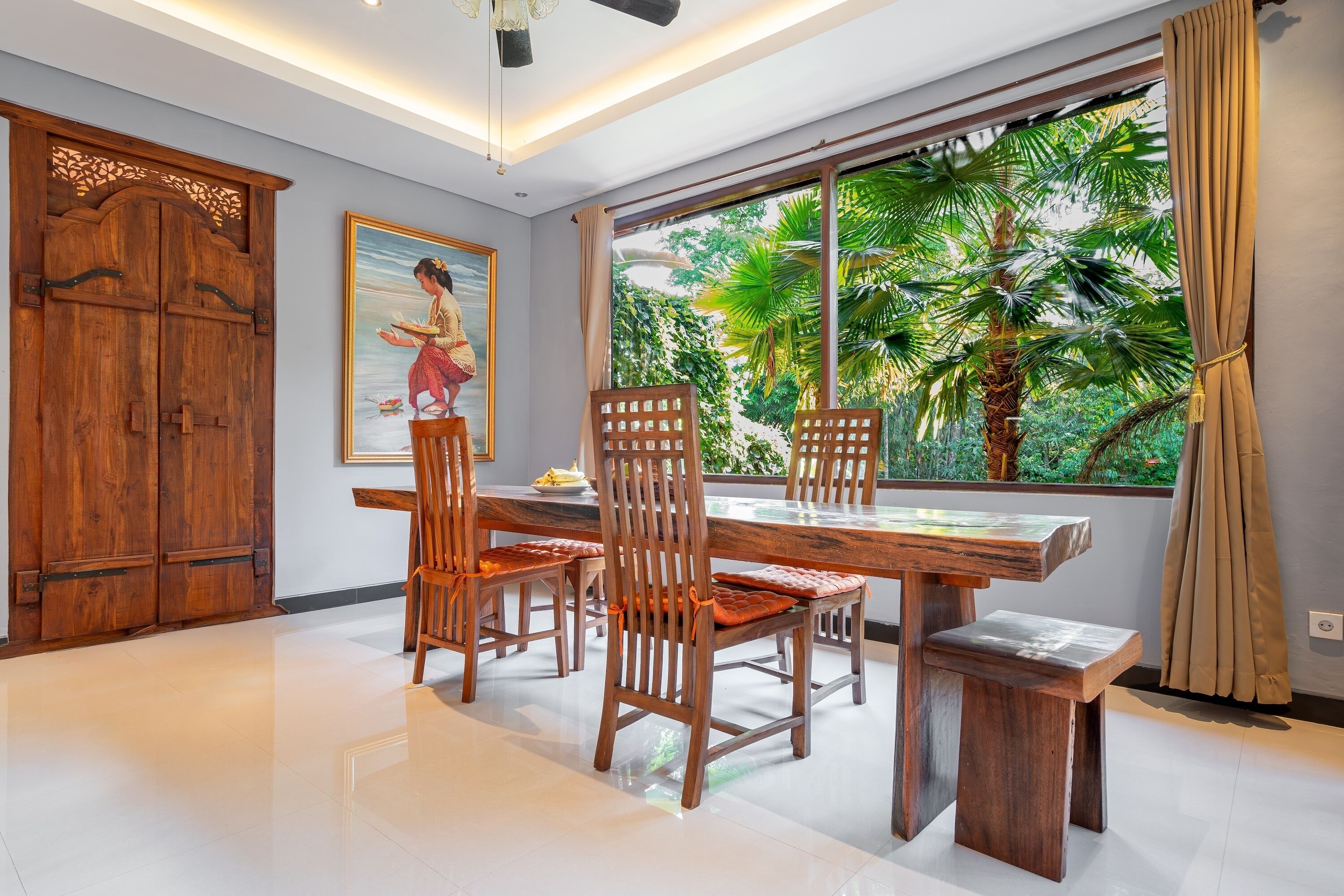 A dining area with a wooden table and chairs, a large window with a view of palm trees, and a traditional wooden door.