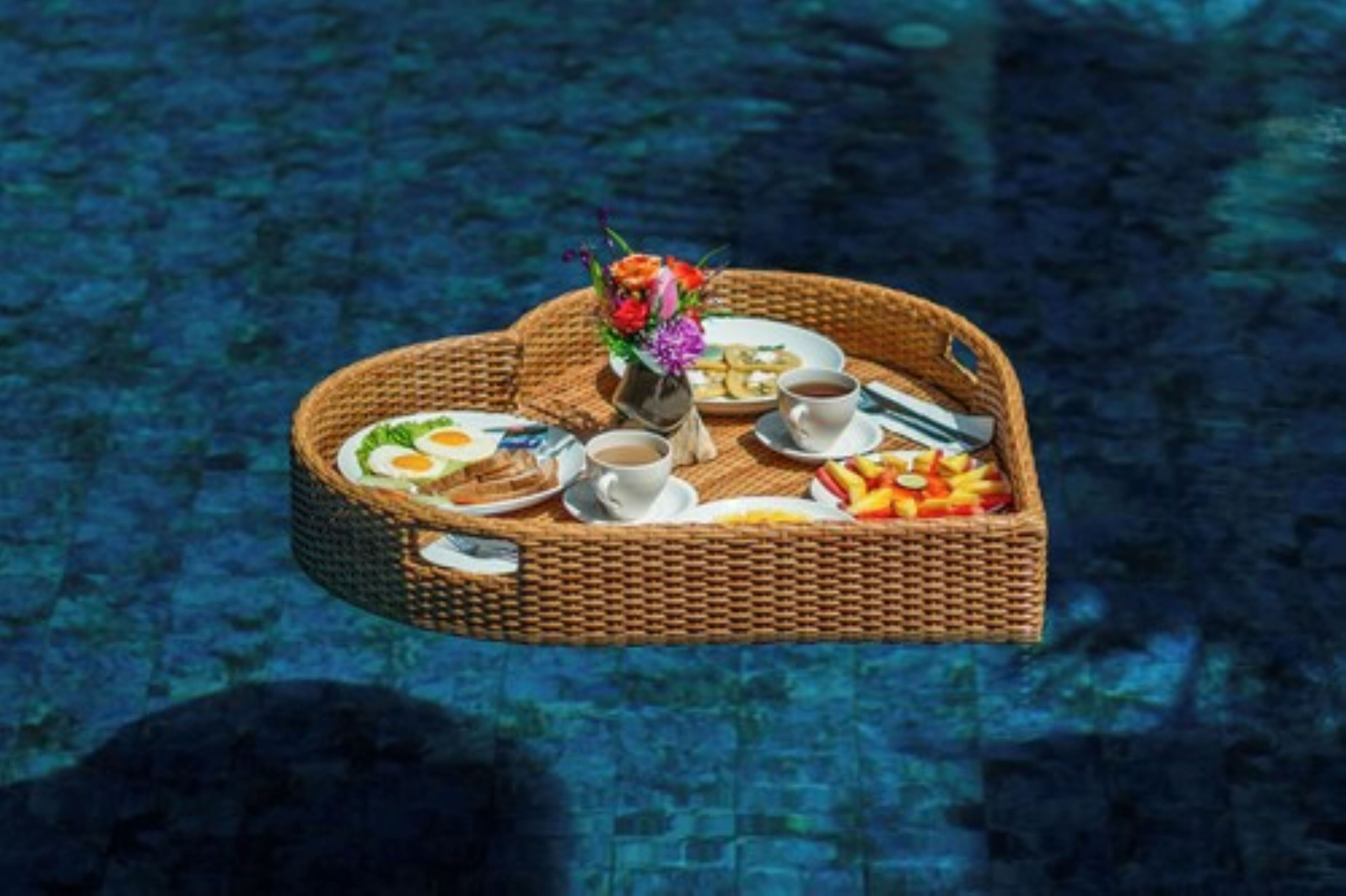 A floating breakfast setup with a wicker tray containing croissants, coffee, and fruit, set against a pool backdrop.