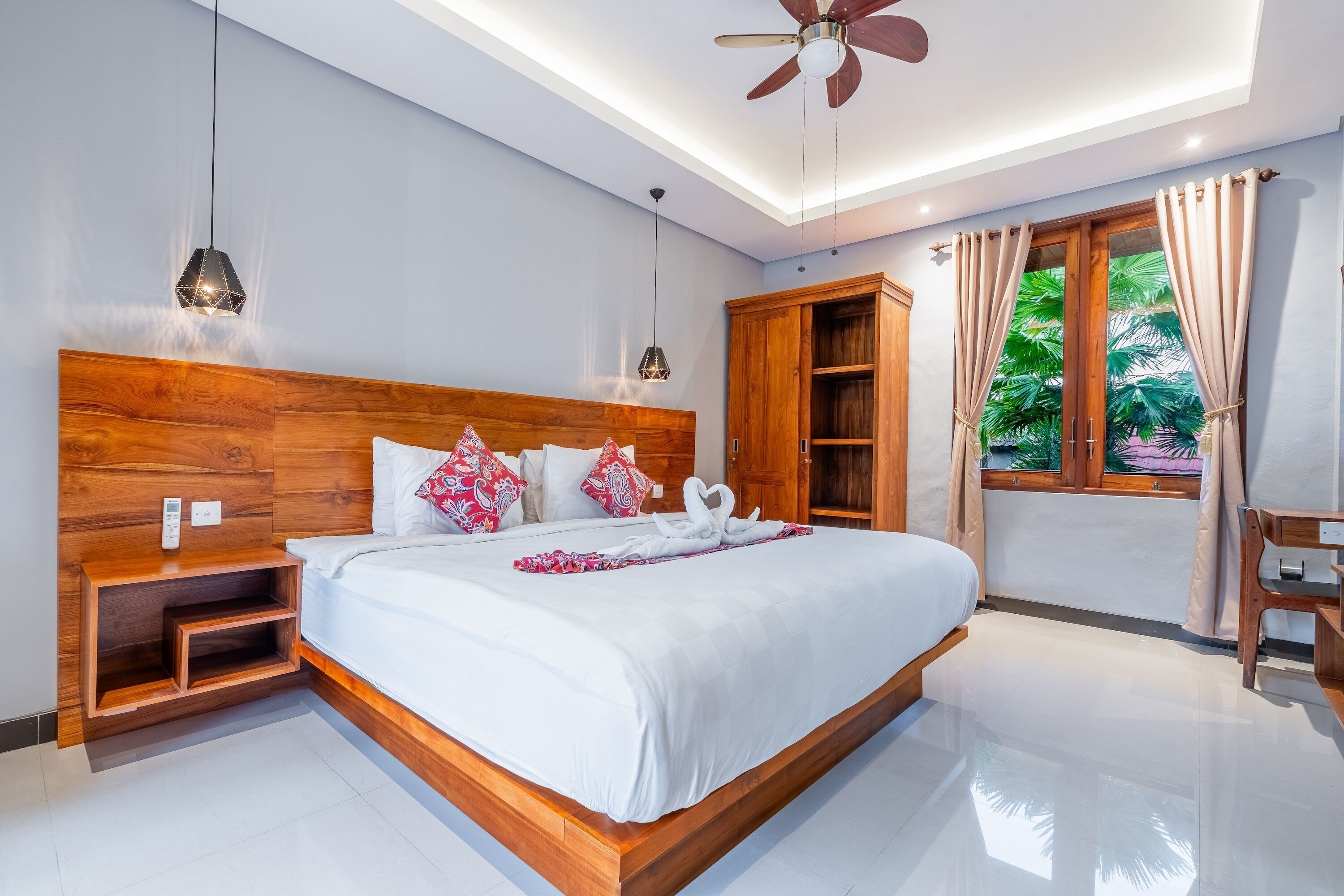 A modern bedroom with a wooden headboard, a bed with white bedding, a bedside table, a nightstand, a ceiling fan, a window with curtains, and a view of greenery outside.