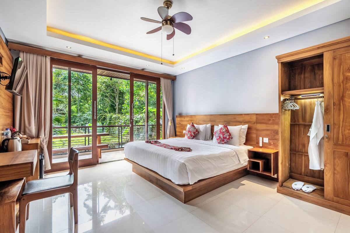 Une chambre dotée d’un grand lit, d’une tête de lit en bois, d’un ventilateur de plafond, d’une armoire en bois et offrant une vue sur la verdure grâce à de grandes fenêtres.