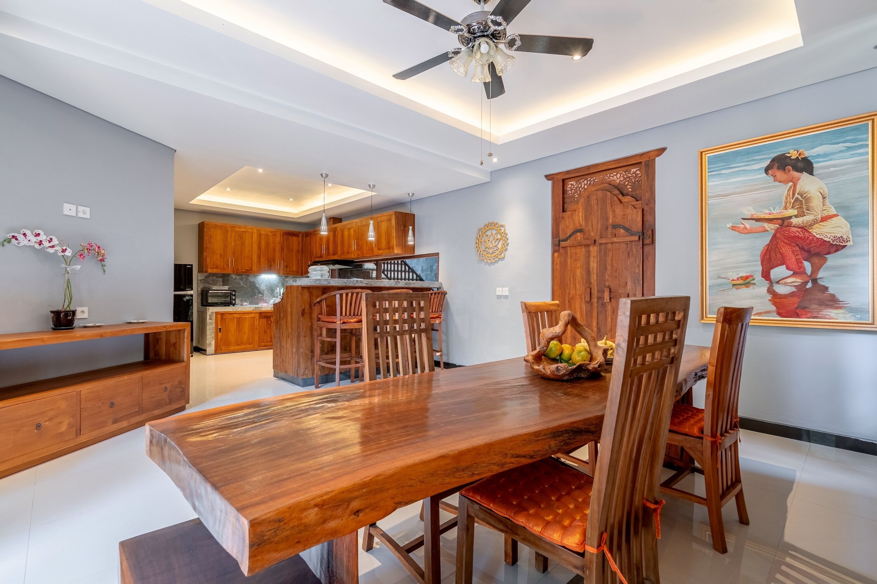 Uma área de refeições moderna com mesa e cadeiras de madeira, um ventilador de teto e uma cozinha ao fundo.