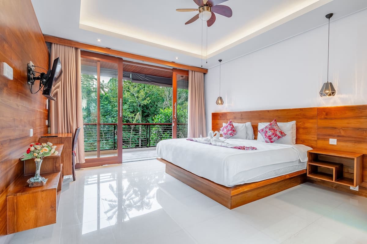 Une chambre moderne avec un grand lit, une tête de lit en bois, un ventilateur de plafond et une vue sur la verdure grâce à une porte-fenêtre coulissante.