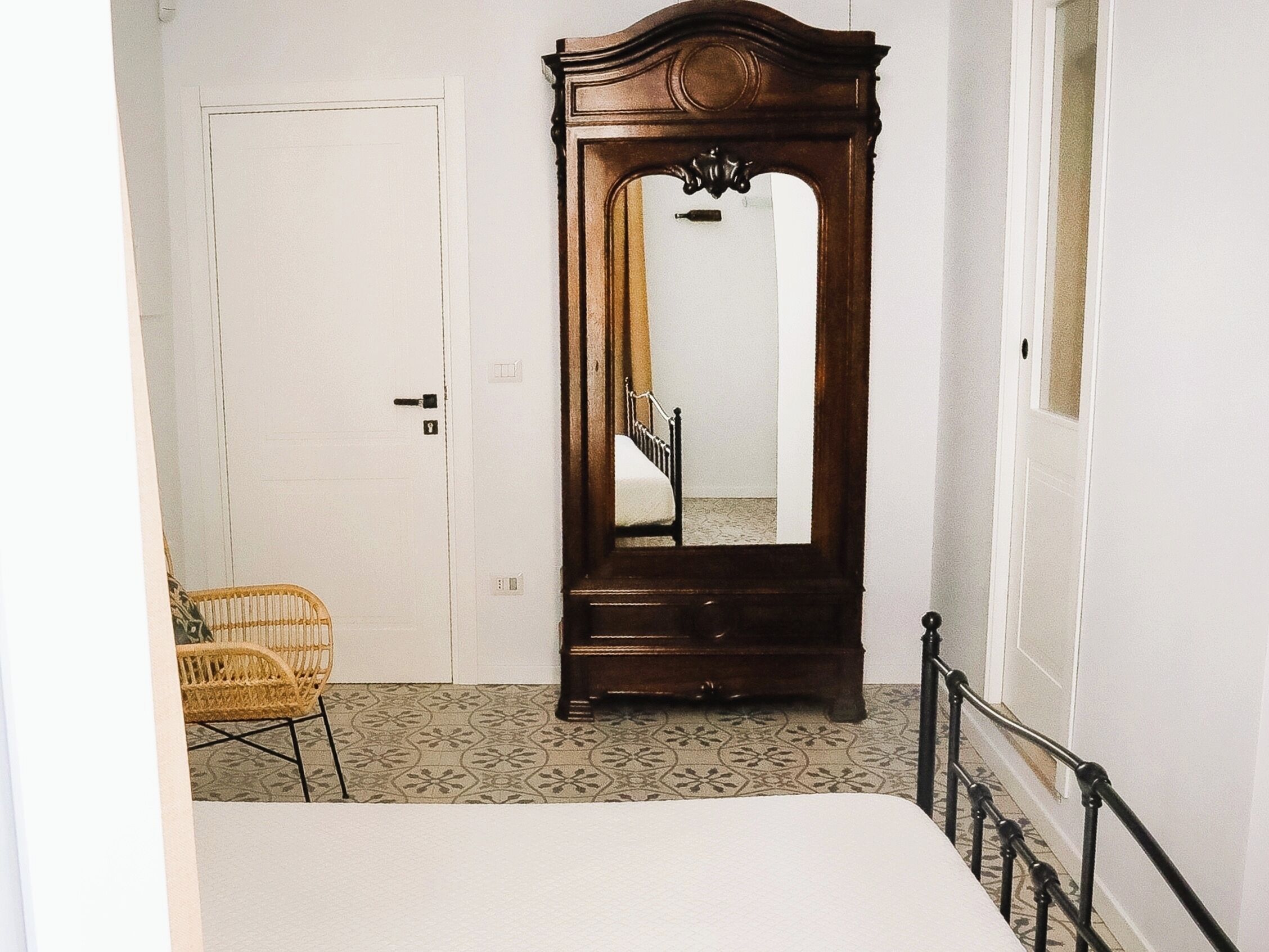A room with a bed, a wooden wardrobe, a wicker chair, and a patterned floor.