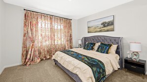 Een slaapkamer met een groot bed, bloemen gordijnen, een landschap schilderij en een nachtkastje met een lamp.