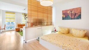 A modern bedroom with a bed, a window with blinds, a painting, and a small table with a lamp.