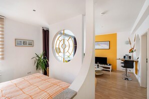 A modern hotel room with a bed, a round window, a TV, a dining area, and a plant.