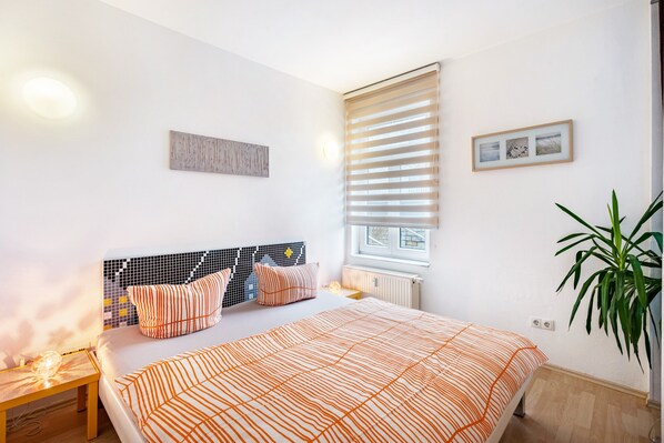 A bedroom with a bed, a nightstand, a window with blinds, a radiator, and a potted plant.