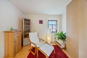 Un espace de vie compact comprenant un meuble en bois, une chaise blanche rembourrée, un tapis rouge, une plante en pot et une fenêtre donnant sur un beau paysage.