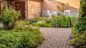 Un edificio de piedra con una puerta de madera, un jardín empedrado con plantas y dos sillas blancas cerca del edificio.