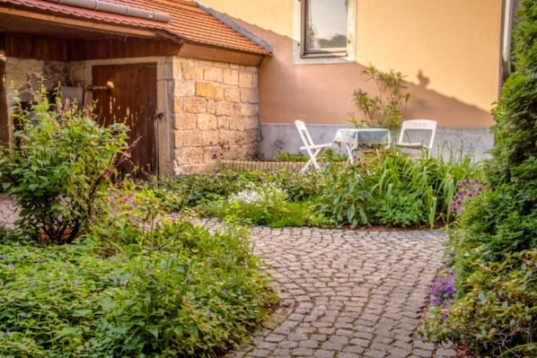 Un bâtiment en pierre avec une porte en bois, un jardin aménagé avec des plantes et deux chaises blanches près du bâtiment.